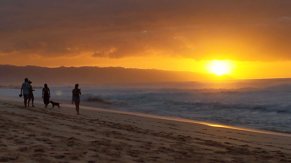 A photo of the sunset taken from the beach at pipeline showing Kaena Point and people walking on.